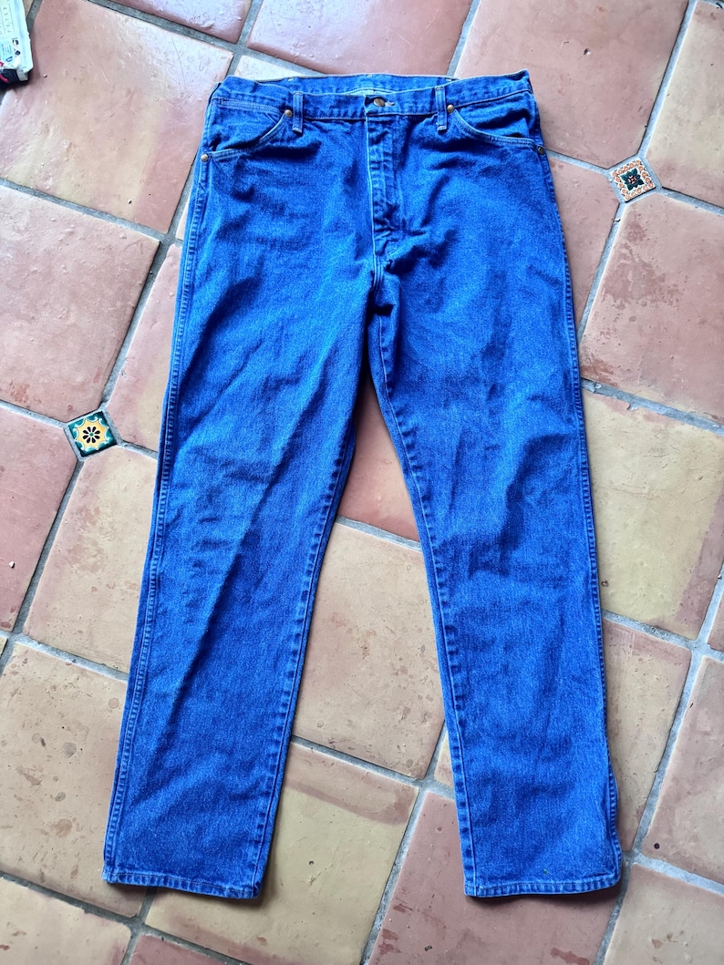 Puede incluir: Un par de vaqueros de mezclilla azul medio con un dise&ntilde;o cl&aacute;sico de cinco bolsillos, cierre de botones y remaches de color cobre en las esquinas de los bolsillos. Los vaqueros tienen un corte recto con costuras visibles en las piernas.