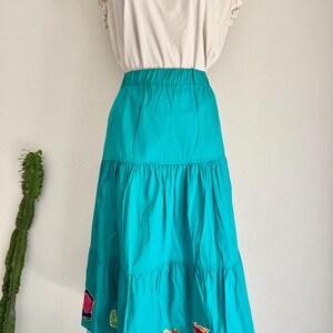 May include: A two-piece outfit featuring a cream-colored top with ruffled sleeves and a turquoise tiered skirt. The skirt has colorful appliqués near the hem. The outfit is displayed against a white wall with a cactus in the background.