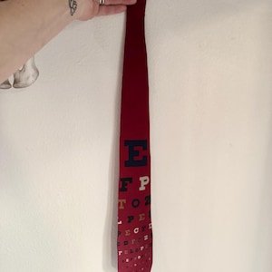 May include: A burgundy necktie featuring a classic eye chart design. The tie has a solid burgundy color at the top and transitions to the eye chart with black, white, and gold lettering. The tie is held up by a hand.