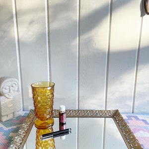 May include: A gold mirrored tray with an ornate design and feet. The tray has a glass tumbler and a small bottle on top of it.