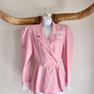 May include: A light pink blazer featuring long sleeves with white lace cuffs, a peplum waist, and a double-breasted front with two white buttons. The garment includes a notched lapel collar and a small chest pocket accented with white lace trim.