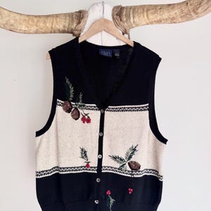 May include: A black and cream knit button-down vest featuring a V-neckline and a ribbed hem. The garment displays an embroidered pattern of brown pinecones, green pine needles, and small red berries across the front panels and chest area.