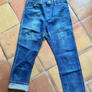 May include: Blue denim jeans with a button fly and a classic five-pocket design. The jeans have a faded wash and are cuffed at the ankles, revealing the lighter denim. The jeans are laid flat on a tiled surface.