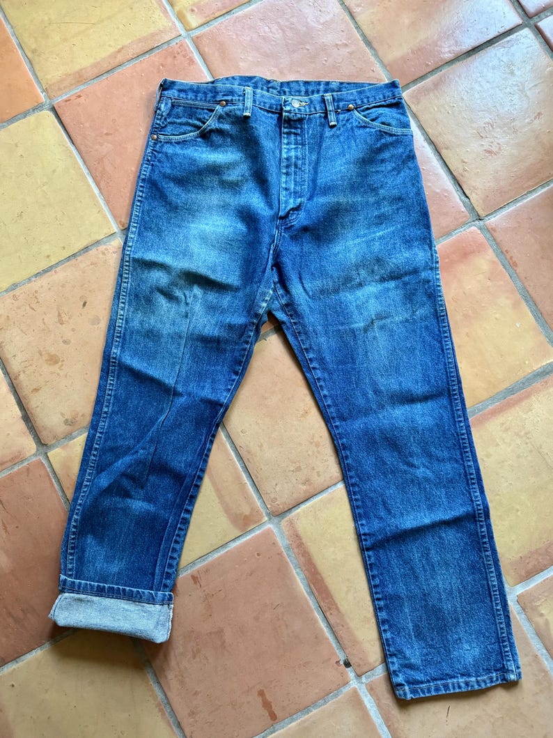 May include: Blue denim jeans with a button fly and a classic five-pocket design. The jeans have a faded wash and are cuffed at the ankles, revealing the lighter denim. The jeans are laid flat on a tiled surface.