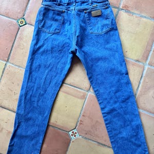 Puede incluir: Unos vaqueros de mezclilla azul con un dise&ntilde;o cl&aacute;sico de cinco bolsillos, cierre de bot&oacute;n y cremallera, trabillas para cintur&oacute;n y un parche de cuero marr&oacute;n en el bolsillo trasero derecho con la marca Wrangler visible en la tela.