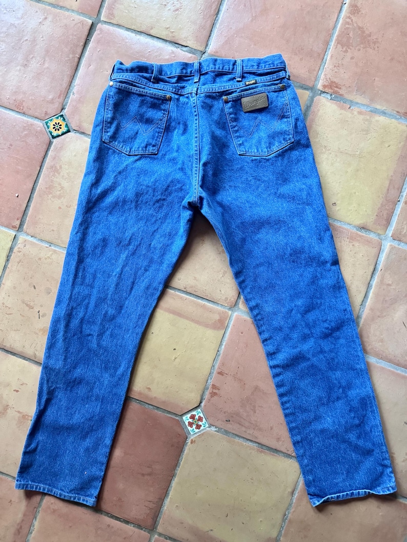 Puede incluir: Unos vaqueros de mezclilla azul con un dise&ntilde;o cl&aacute;sico de cinco bolsillos, cierre de bot&oacute;n y cremallera, trabillas para cintur&oacute;n y un parche de cuero marr&oacute;n en el bolsillo trasero derecho con la marca Wrangler visible en la tela.