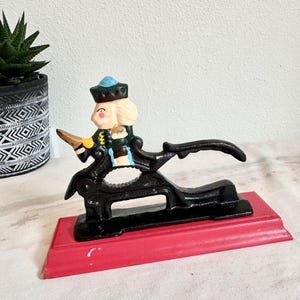 May include: A vintage cast iron nutcracker featuring a soldier figure with a blue hat and blonde hair. The black nutcracker is mounted on a red wooden base. The soldier is dressed in a green and black uniform.