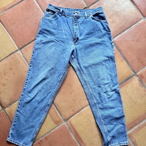 May include: Light blue denim jeans with a classic five-pocket design. The jeans have a button and zipper closure, belt loops, and a straight leg cut. The fabric shows subtle fading and wear, suggesting a vintage or pre-worn style.