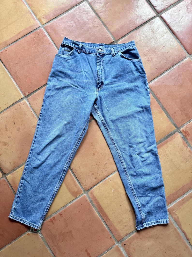 May include: Light blue denim jeans with a classic five-pocket design. The jeans have a button and zipper closure, belt loops, and a straight leg cut. The fabric shows subtle fading and wear, suggesting a vintage or pre-worn style.