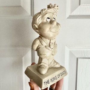 May include: A vintage figurine of a man wearing a crown, suit, and tie, standing on a base that reads "THE KING OF DADS." The figurine is a cream color and has a smiling expression. The base is a light beige color.