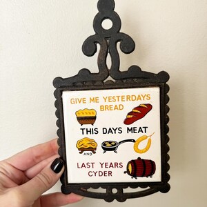 May include: A cast iron trivet with a white ceramic tile insert. The tile has a colorful illustration and the text "Give me yesterdays bread, this days meat and last years cyder".