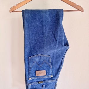 May include: Blue denim jeans hanging on a wooden hanger. The jeans have a classic five-pocket design with a brown leather patch on the back pocket that says "Wrangler". The jeans are a medium wash.