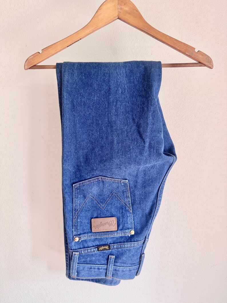 May include: Blue denim jeans hanging on a wooden hanger. The jeans have a classic five-pocket design with a brown leather patch on the back pocket that says "Wrangler". The jeans are a medium wash.