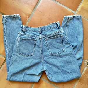 May include: Light blue denim jeans with a classic five-pocket design. The jeans are laid flat, showcasing the back pockets, belt loops, and button closure. The denim has a faded appearance, suggesting a vintage or worn look.
