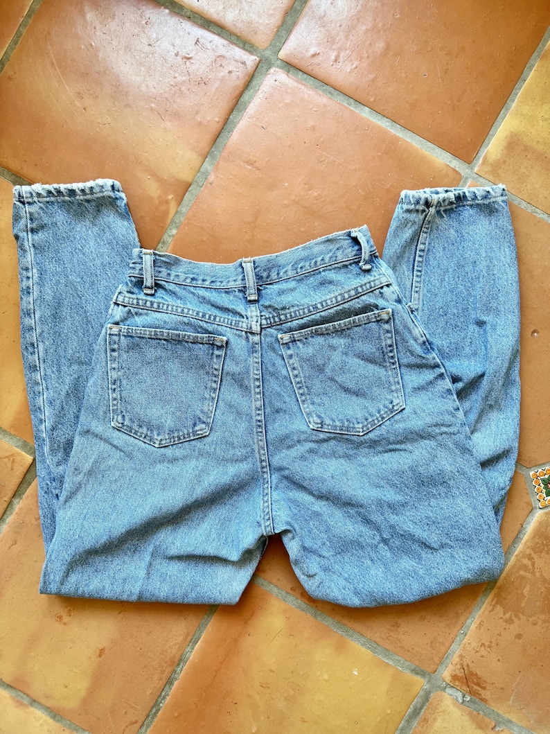 May include: Light blue denim jeans with a classic five-pocket design. The jeans are laid flat, showcasing the back pockets, belt loops, and button closure. The denim has a faded appearance, suggesting a vintage or worn look.