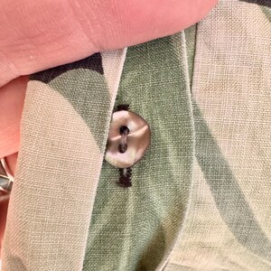 Peut inclure: Gros plan sur une chemise boutonn&eacute;e. Le bouton est de couleur claire et iris&eacute;e, et le tissu de la chemise est vert clair avec un motif subtil. La chemise est partiellement d&eacute;boutonn&eacute;e, r&eacute;v&eacute;lant la boutonni&egrave;re.