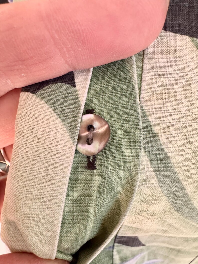 Peut inclure: Gros plan sur une chemise boutonn&eacute;e. Le bouton est de couleur claire et iris&eacute;e, et le tissu de la chemise est vert clair avec un motif subtil. La chemise est partiellement d&eacute;boutonn&eacute;e, r&eacute;v&eacute;lant la boutonni&egrave;re.