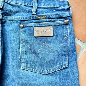 May include: Close-up of blue denim jeans with a back pocket. The pocket features a stitched design and a brown leather patch with the "Wrangler" logo. The jeans have a classic design with a button and zipper closure.