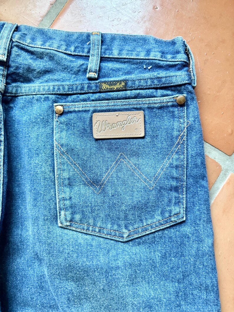 May include: Close-up of blue denim jeans with a back pocket. The pocket features a stitched design and a brown leather patch with the "Wrangler" logo. The jeans have a classic design with a button and zipper closure.