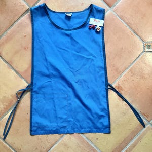 May include: A bright blue apron with a dark blue trim and ties. It features a small pocket on the front and a tag with the word "Pilgrim" and a small pin. The apron is made of a smooth fabric.