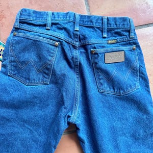 May include: Close-up of the rear of blue denim jeans. The jeans feature two back pockets with a stitched "W" design, a brown leather patch with the "Wrangler" logo, and a belt loop. The jeans are made of a sturdy denim material.