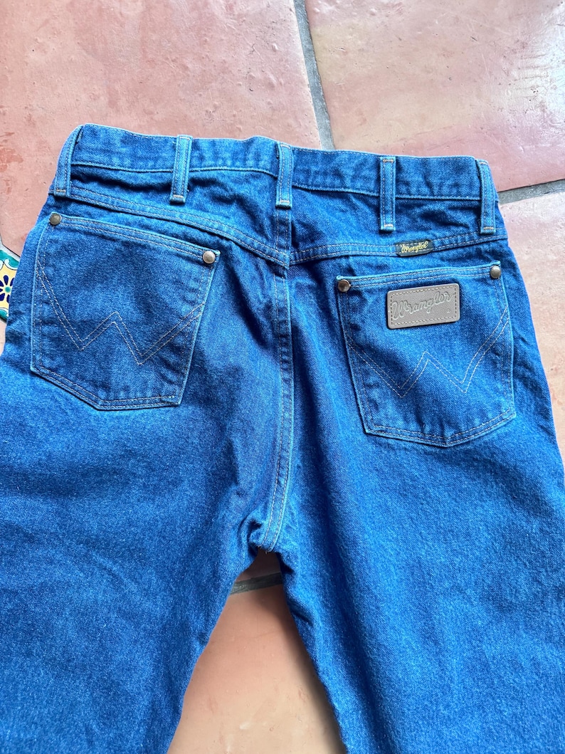May include: Close-up of the rear of blue denim jeans. The jeans feature two back pockets with a stitched "W" design, a brown leather patch with the "Wrangler" logo, and a belt loop. The jeans are made of a sturdy denim material.