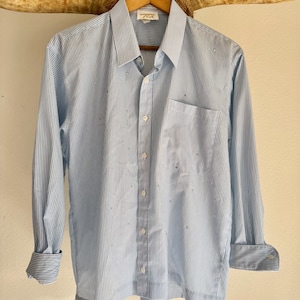 May include: Light blue and white striped button-down shirt with long sleeves. The shirt has a chest pocket and is hanging on a wooden hanger. The shirt has small, scattered embellishments.