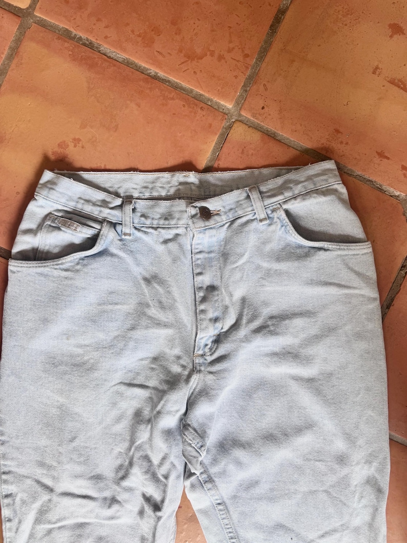 May include: Light blue denim jeans with a button closure and classic five-pocket design. The jeans are made of a light-wash denim fabric and have a straight leg cut. The jeans are shown on a terracotta tile surface.