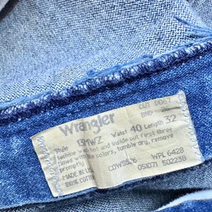 May include: Close-up of blue denim jeans with a Wrangler label. The label indicates a waist size of 40 inches and a length of 32 inches. The jeans are made of 100% cotton.
