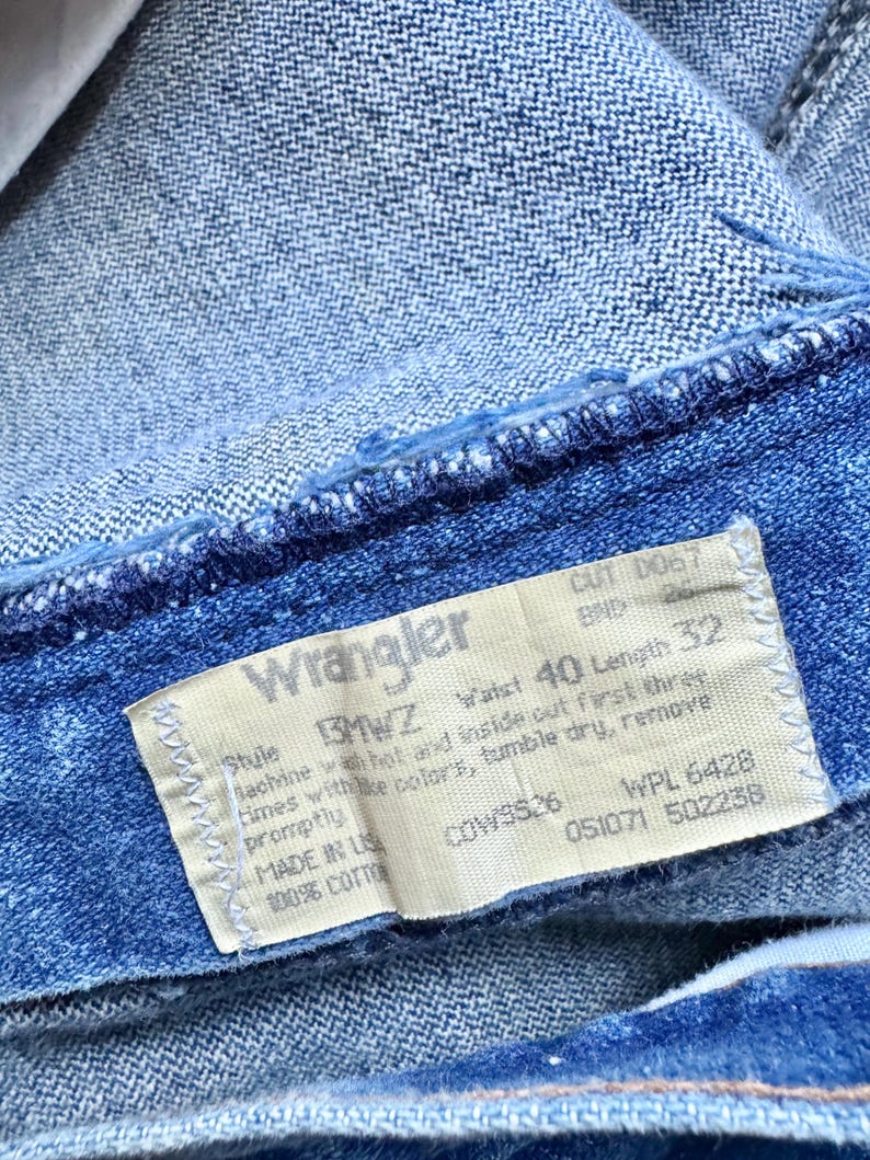 May include: Close-up of blue denim jeans with a Wrangler label. The label indicates a waist size of 40 inches and a length of 32 inches. The jeans are made of 100% cotton.