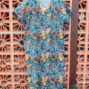 May include: A short-sleeved dress with a V-neck, featuring a vibrant tropical leaf pattern in shades of blue, green, yellow, and red. The dress is hanging on a wooden hanger, showcasing its knee-length design and button-down front.
