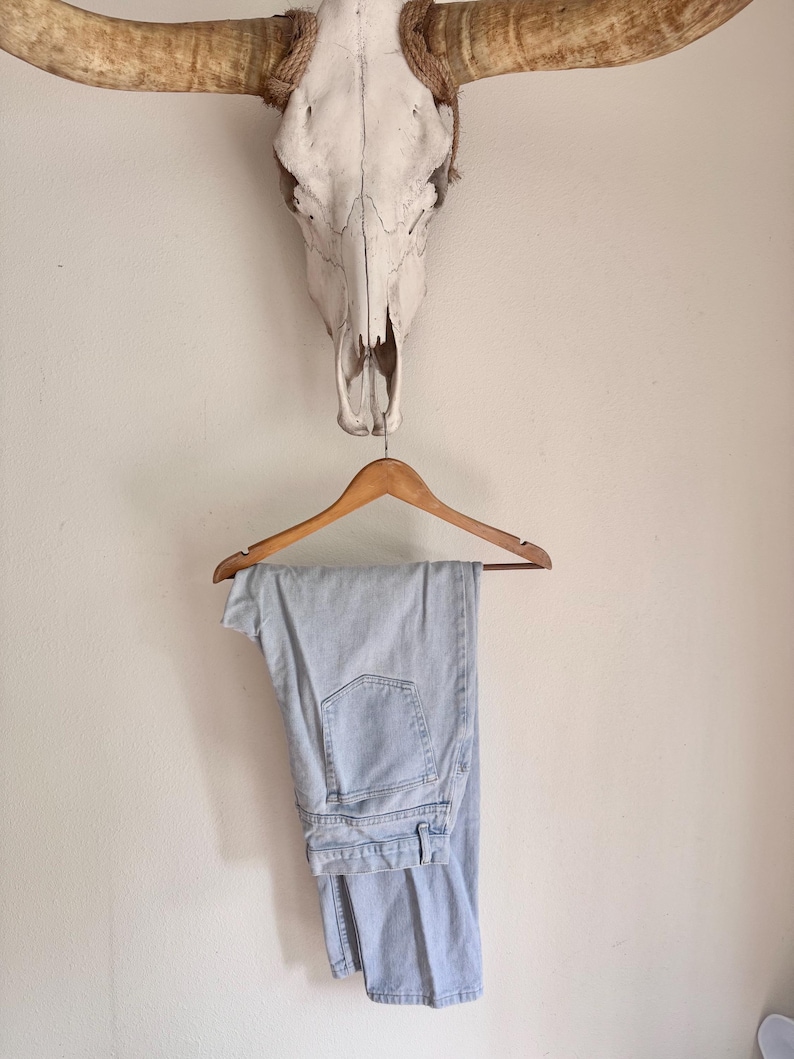 May include: Light blue denim jeans hanging on a wooden hanger. The jeans are folded and display the back pockets and belt loops. A cow skull with large horns is mounted on the wall above the jeans.