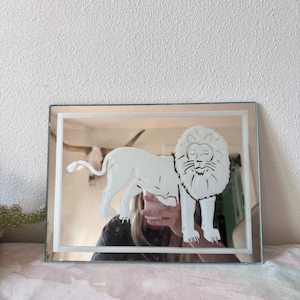 May include: Rectangular mirror with a white lion design etched into the glass. The lion is depicted in a standing pose within a white rectangular border. The mirror has a reflective surface.