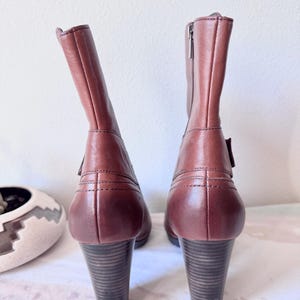 May include: A pair of brown leather ankle boots with stacked heels. The boots have a zipper on the side and a buckle detail. The heels are approximately 3 inches tall.