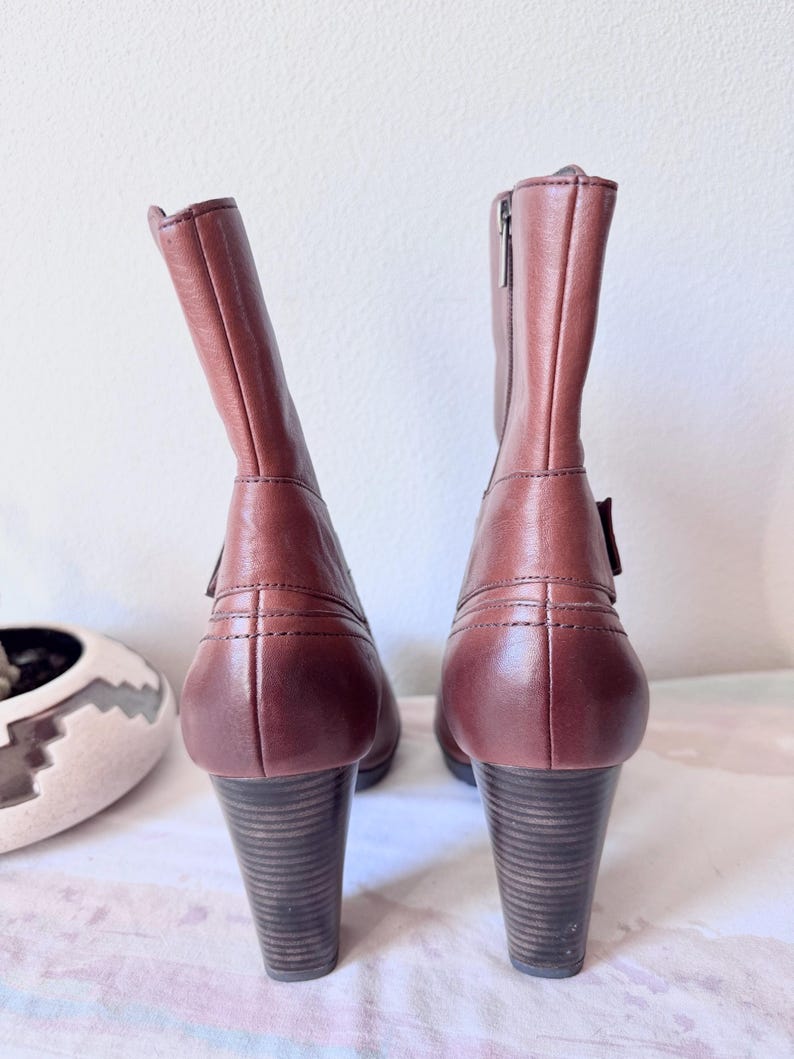 May include: A pair of brown leather ankle boots with stacked heels. The boots have a zipper on the side and a buckle detail. The heels are approximately 3 inches tall.