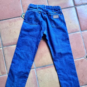 May include: Blue denim jeans with a classic five-pocket design. The jeans feature a Wrangler brand patch on the back and a tag on the back pocket. The jeans are laid flat on a tiled surface.