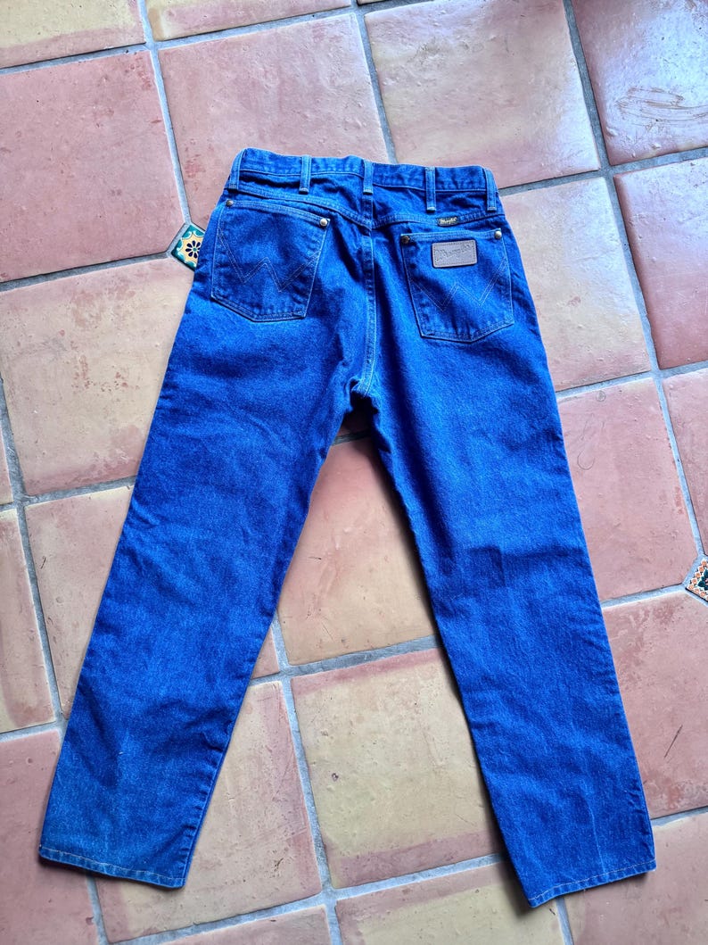 May include: Blue denim jeans with a classic five-pocket design. The jeans feature a Wrangler brand patch on the back and a tag on the back pocket. The jeans are laid flat on a tiled surface.