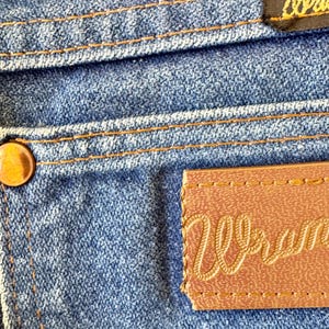 May include: Close-up of blue denim jeans with orange stitching. A brown leather patch with the word "Wrangler" in cursive script is visible, along with a small black tag with the word "Zero" on it.