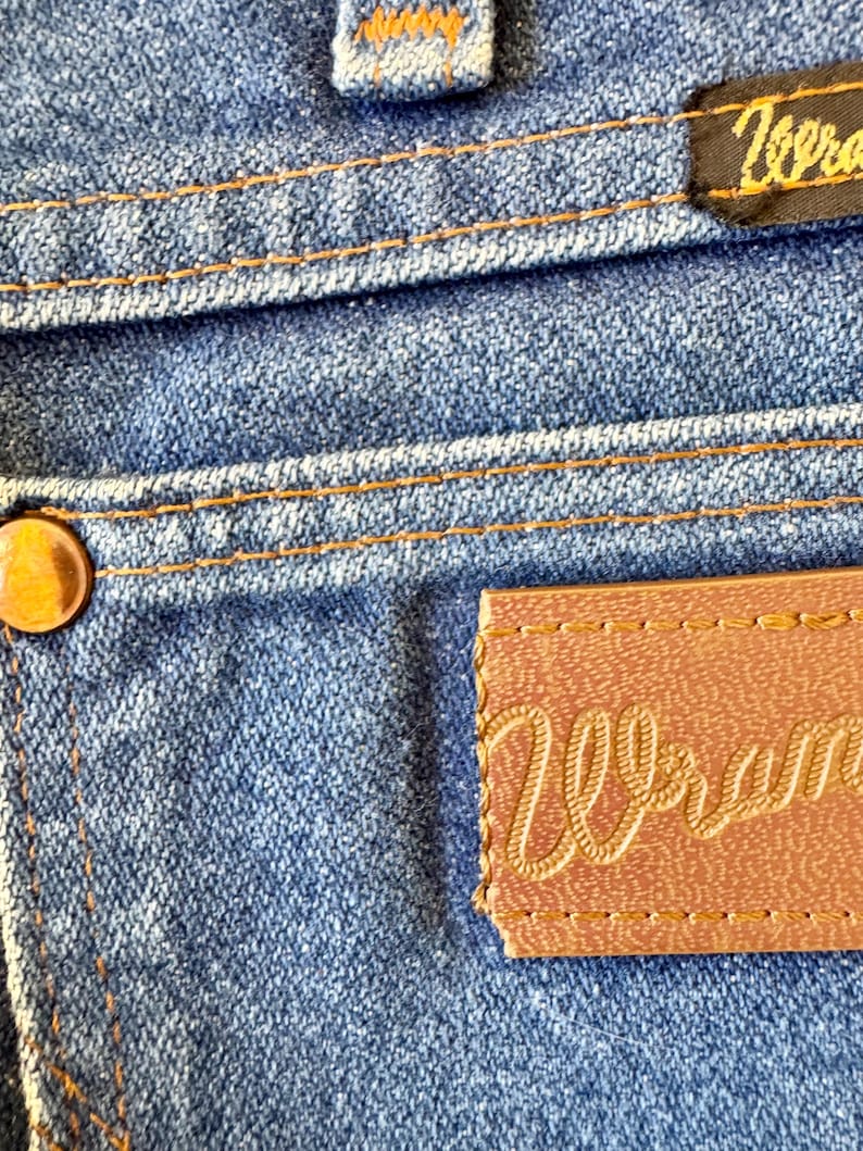 May include: Close-up of blue denim jeans with orange stitching. A brown leather patch with the word "Wrangler" in cursive script is visible, along with a small black tag with the word "Zero" on it.