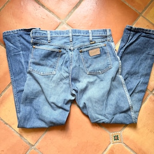 K&ouml;nnte beinhalten: Hellblaue Jeans mit klassischem F&uuml;nf-Taschen-Design. Die Jeans haben einen braunen Lederaufn&auml;her mit dem Markennamen und einen Metallknopfverschluss. Die Ges&auml;&szlig;taschen haben ein klassisches Nahtdesign. Die Jeans liegen flach auf einer gefliesten Oberfl&auml;che.