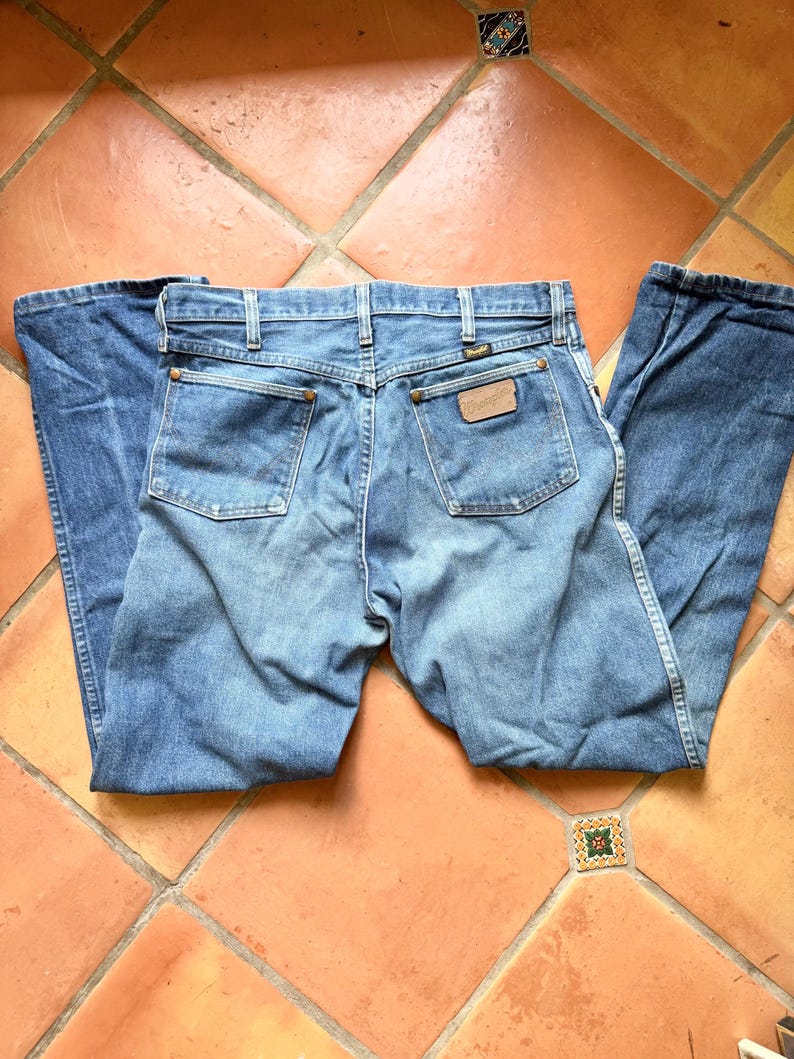 K&ouml;nnte beinhalten: Hellblaue Jeans mit klassischem F&uuml;nf-Taschen-Design. Die Jeans haben einen braunen Lederaufn&auml;her mit dem Markennamen und einen Metallknopfverschluss. Die Ges&auml;&szlig;taschen haben ein klassisches Nahtdesign. Die Jeans liegen flach auf einer gefliesten Oberfl&auml;che.
