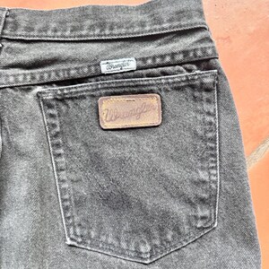 Op de afbeelding: Close-up van een zwarte Wrangler-jeans. De jeans heeft een klassiek ontwerp met vijf zakken, met een klein Wrangler-label boven de achterzak en een grotere leren patch op de achterzak. De jeans is gemaakt van duurzaam denim materiaal.