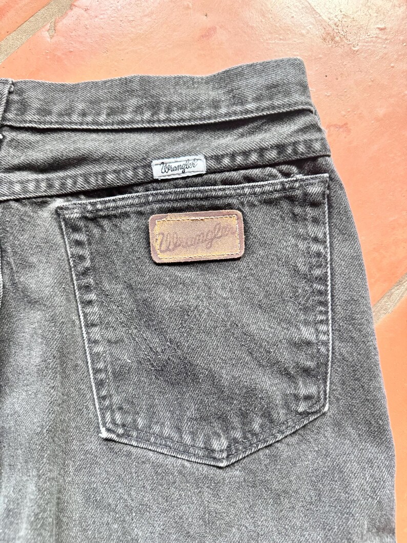 Op de afbeelding: Close-up van een zwarte Wrangler-jeans. De jeans heeft een klassiek ontwerp met vijf zakken, met een klein Wrangler-label boven de achterzak en een grotere leren patch op de achterzak. De jeans is gemaakt van duurzaam denim materiaal.