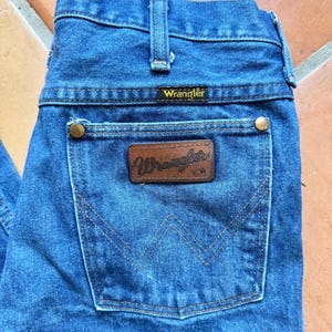 Puede incluir: Primer plano de unos vaqueros Wrangler de mezclilla azul. Los vaqueros presentan un parche de cuero marr&oacute;n con el logotipo de Wrangler y un bolsillo trasero con un dise&ntilde;o cosido. Los vaqueros tienen un dise&ntilde;o cl&aacute;sico de cinco bolsillos con remaches de lat&oacute;n.