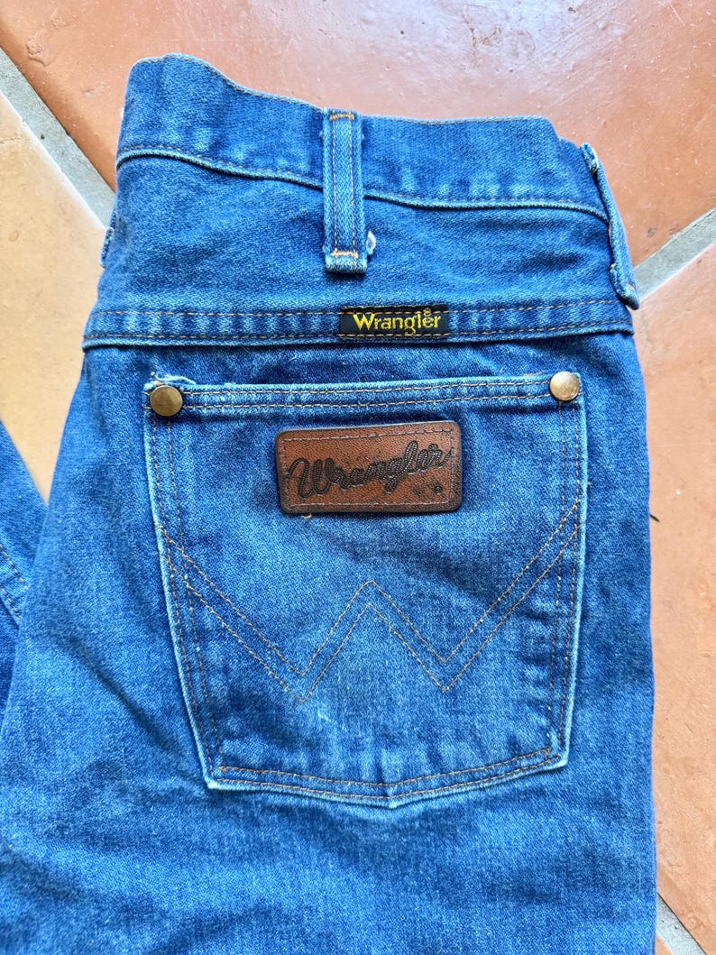 Puede incluir: Primer plano de unos vaqueros Wrangler de mezclilla azul. Los vaqueros presentan un parche de cuero marr&oacute;n con el logotipo de Wrangler y un bolsillo trasero con un dise&ntilde;o cosido. Los vaqueros tienen un dise&ntilde;o cl&aacute;sico de cinco bolsillos con remaches de lat&oacute;n.