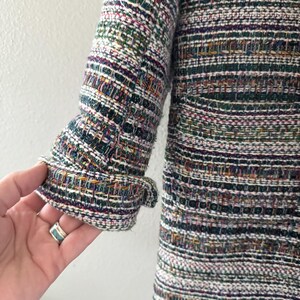 May include: Close-up of a multi-colored tweed jacket sleeve. The sleeve is a mix of green, purple, white, and orange threads. The cuff is folded over, revealing the inside lining. The jacket has a textured appearance.