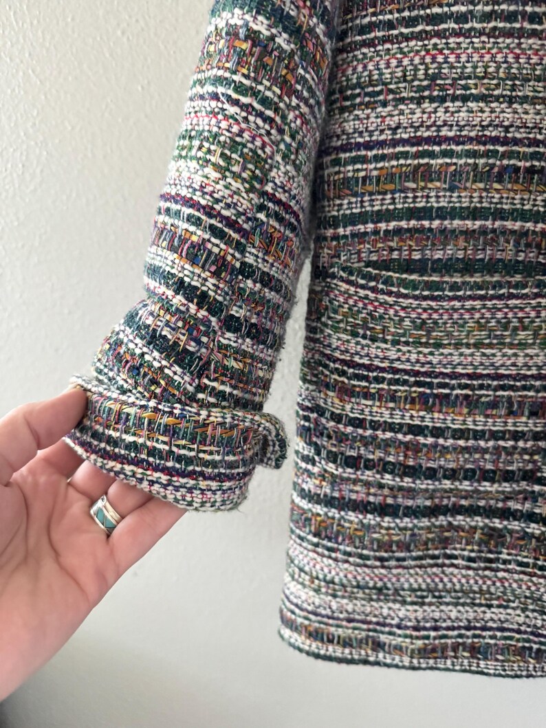 May include: Close-up of a multi-colored tweed jacket sleeve. The sleeve is a mix of green, purple, white, and orange threads. The cuff is folded over, revealing the inside lining. The jacket has a textured appearance.