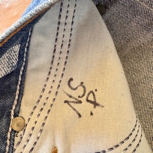 May include: Close-up of the inside of a pair of blue denim jeans. The image shows the white fabric of the pocket with dark blue stitching and the initials "NS D" written in black marker. Rivets and seams are also visible.