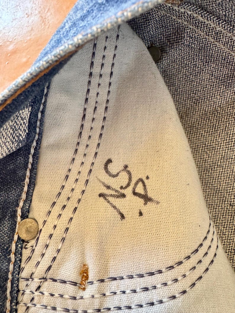 May include: Close-up of the inside of a pair of blue denim jeans. The image shows the white fabric of the pocket with dark blue stitching and the initials "NS D" written in black marker. Rivets and seams are also visible.