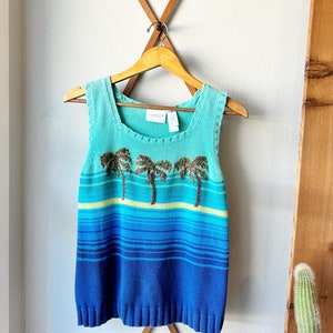 May include: A blue and green striped sleeveless sweater with palm trees embroidered on the front. The sweater is hanging on a wooden hanger.